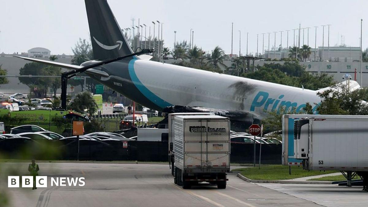 Amazon cargo plane wreckage rests beside an airport service road, surrounded by trucks and emergency vehicles.