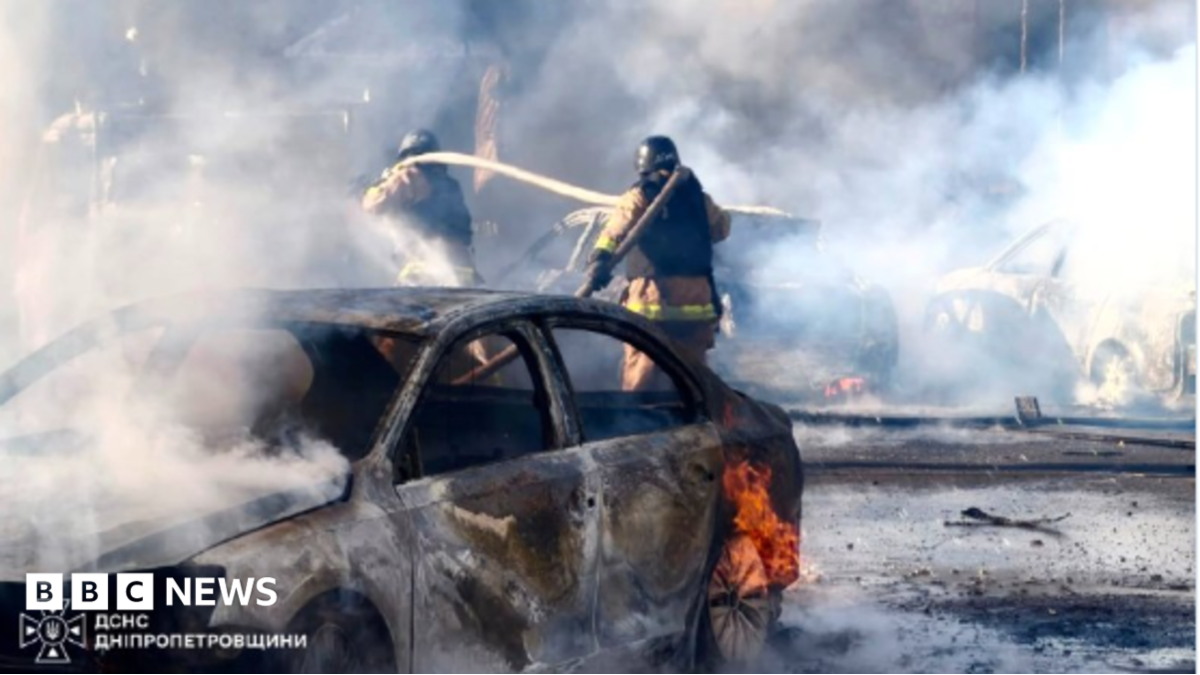 Firefighters spray water near a burning, smoke-filled car amid extensive fire damage.