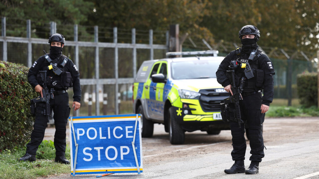 Two armed officers stand beside a police stop sign and marked police vehicle on a roadside.
