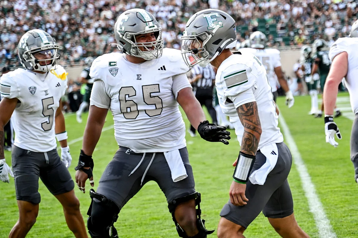 Three football players in white jerseys and silver helmets stand on a field during a game.