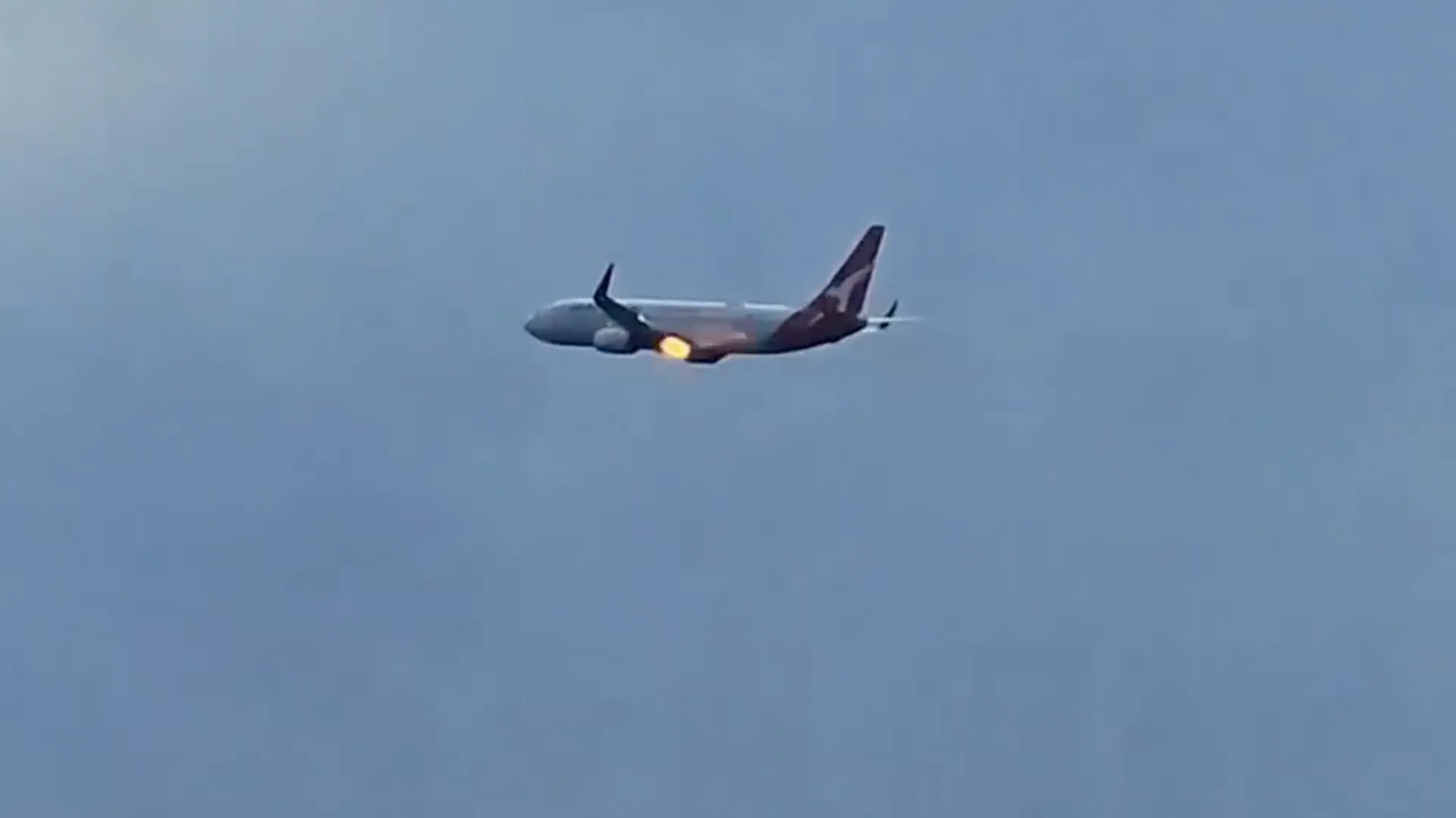 Passenger aircraft in flight with bright flames visible near one engine against a blue sky