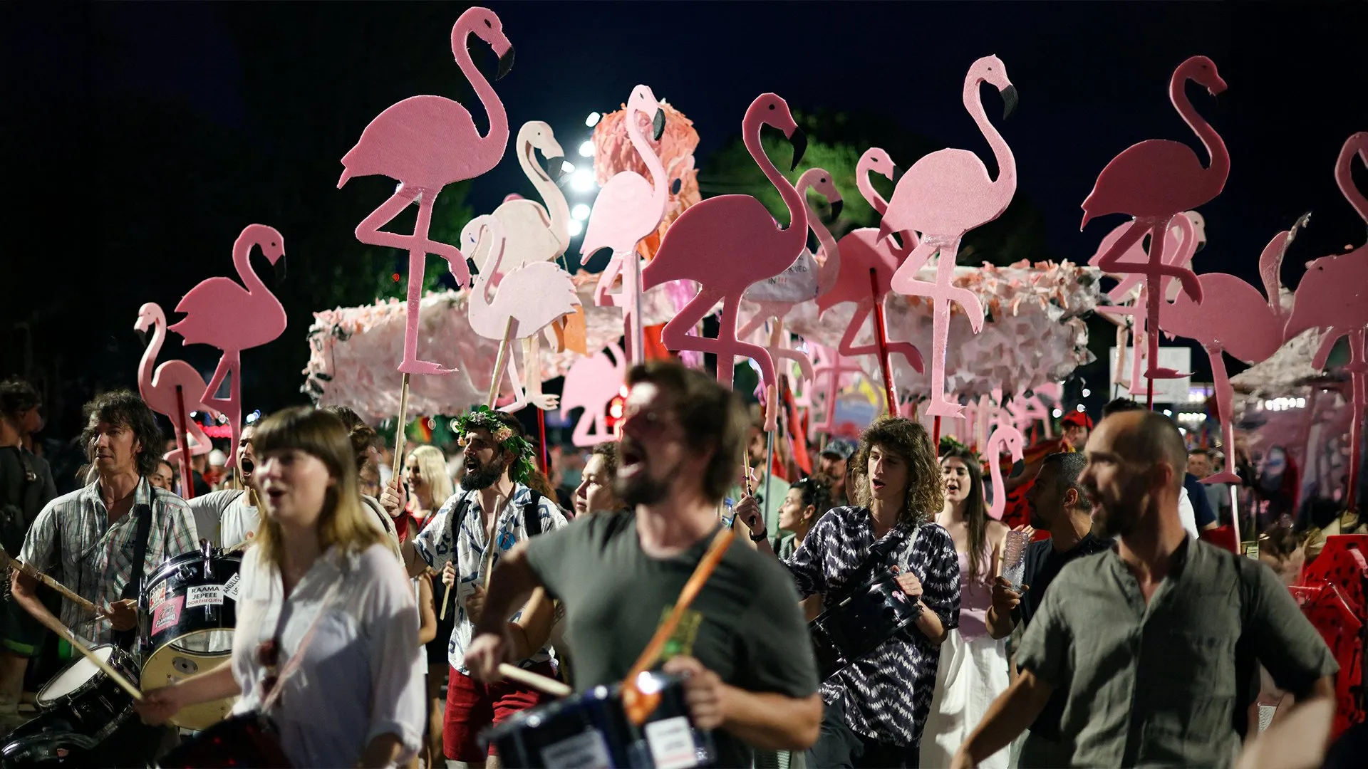 Crowd carrying large pink flamingo signs and playing drums during a nighttime demonstration.