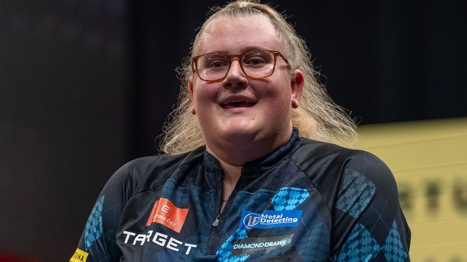 Female darts player wearing glasses and a sponsored dark jersey, smiling during an event