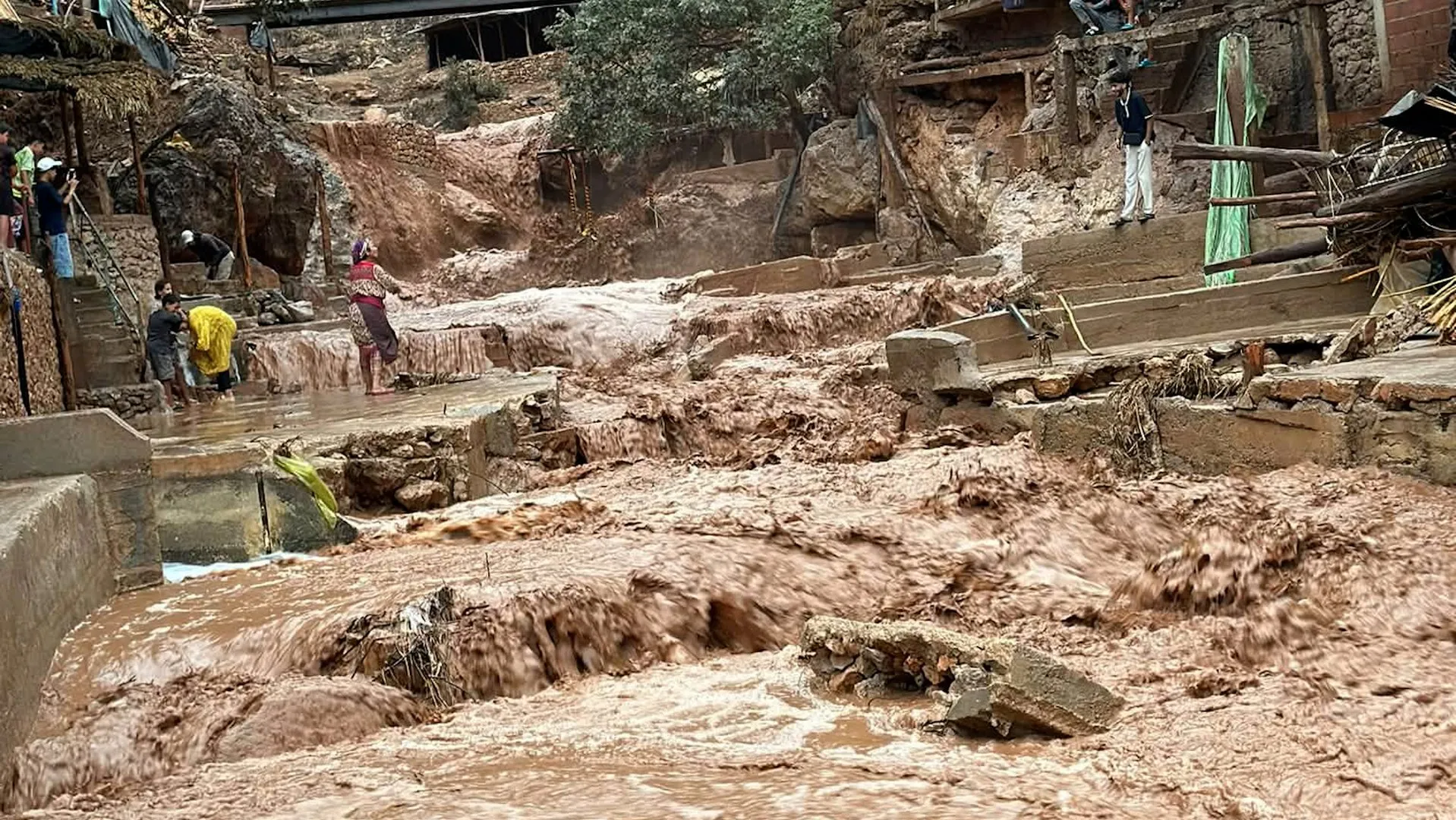 Mud-filled floodwaters rush through a rocky village settlement as residents navigate damaged paths and structures.