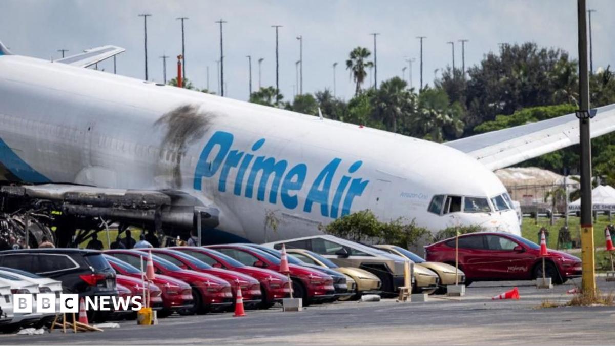 Damaged Prime Air cargo plane beside parked cars and traffic cones at an airport.