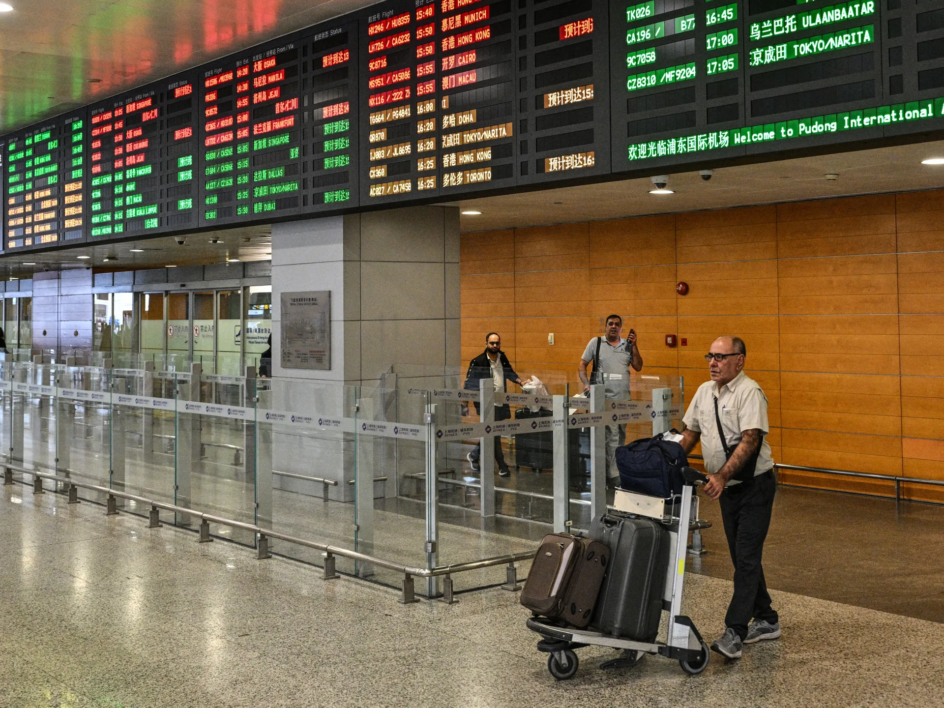 A traveler pushes a luggage cart beneath a large airport flight information board.