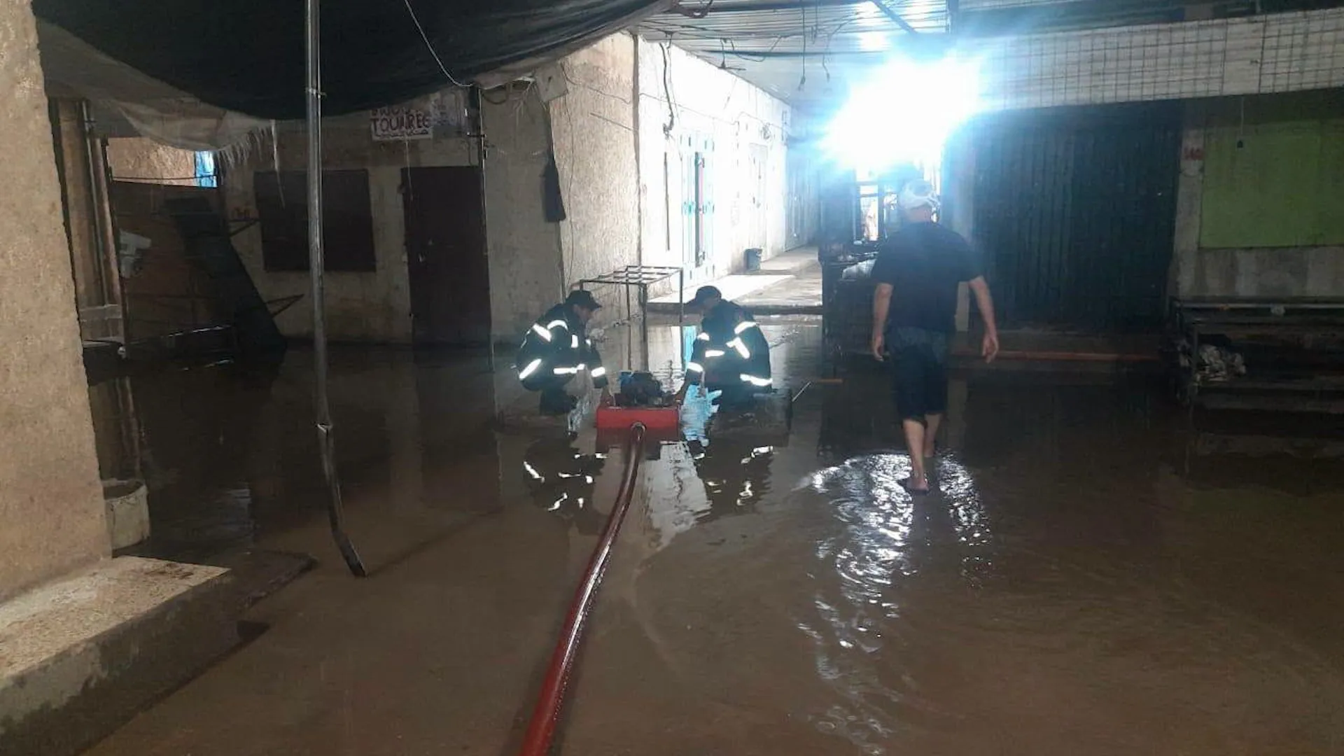 Two responders work beside a hose in a flooded covered passage as a man wades through water.