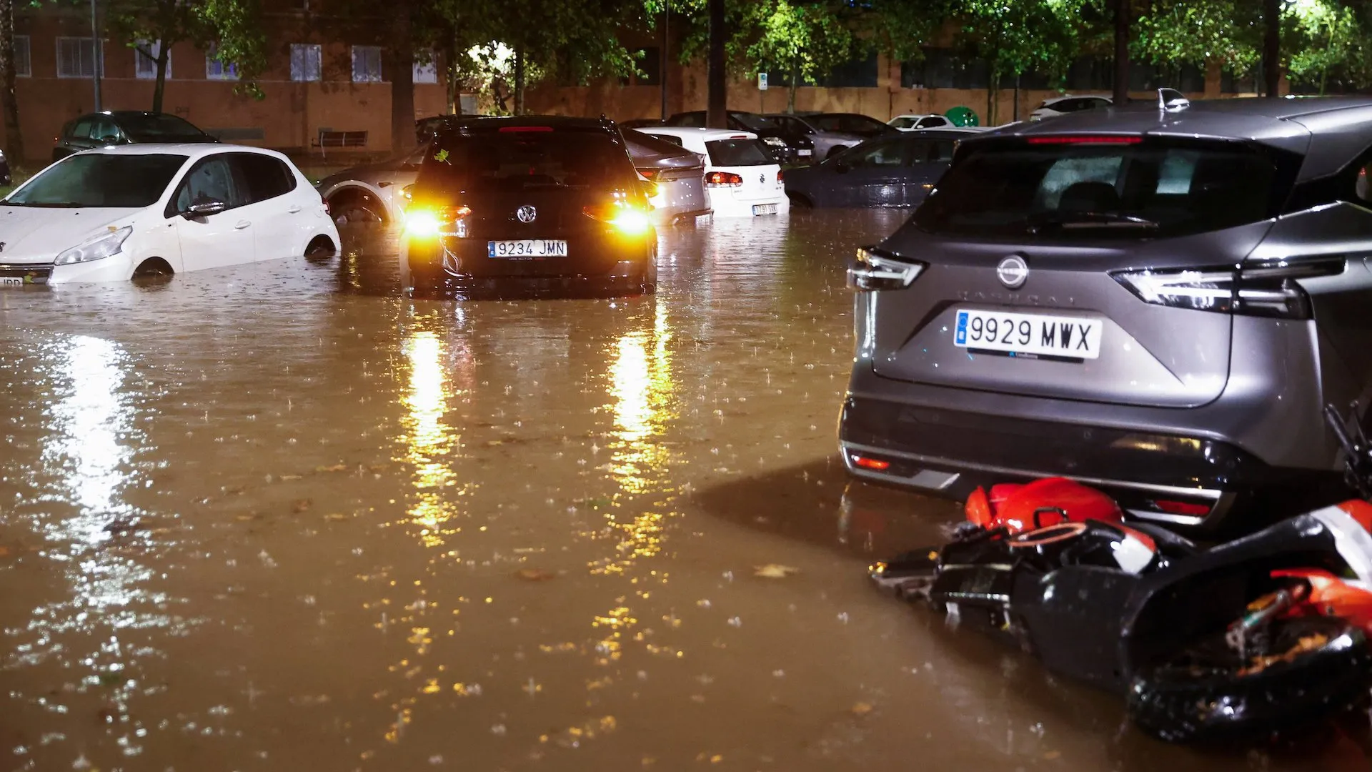 Floodwater surrounds parked cars at night, reflecting headlights and streetlights in a residential area.