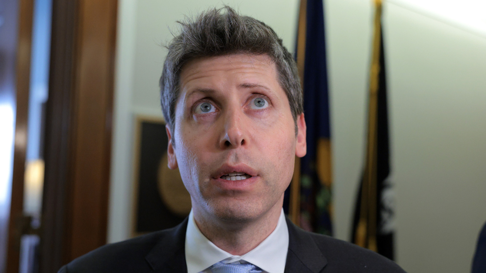 A man in a dark suit and tie looks upward with his mouth slightly open.