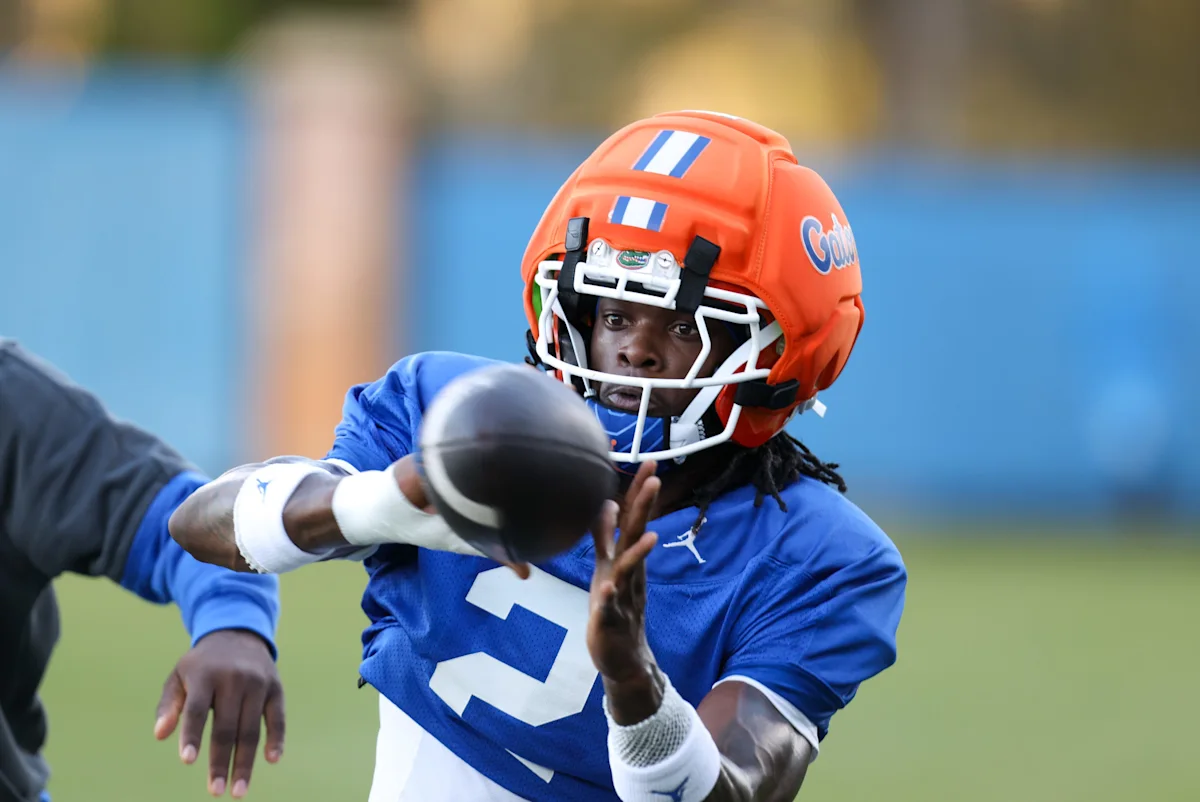 Football player in blue jersey and orange helmet reaches toward a football during practice