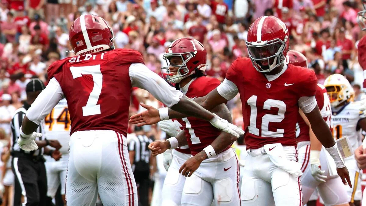 Three Alabama football players in crimson uniforms celebrate during a game as opponents and fans fill the background.