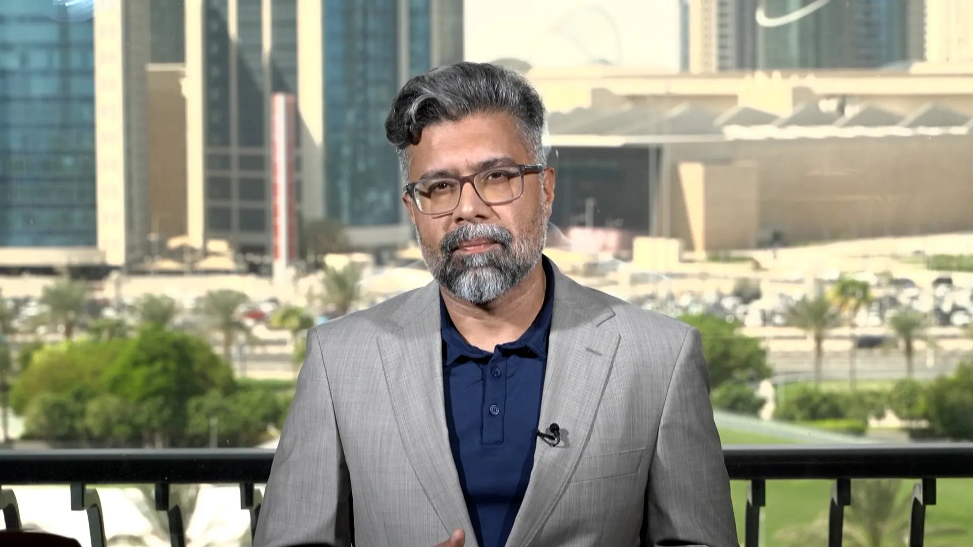 A bespectacled man in a gray suit stands before modern buildings and greenery.