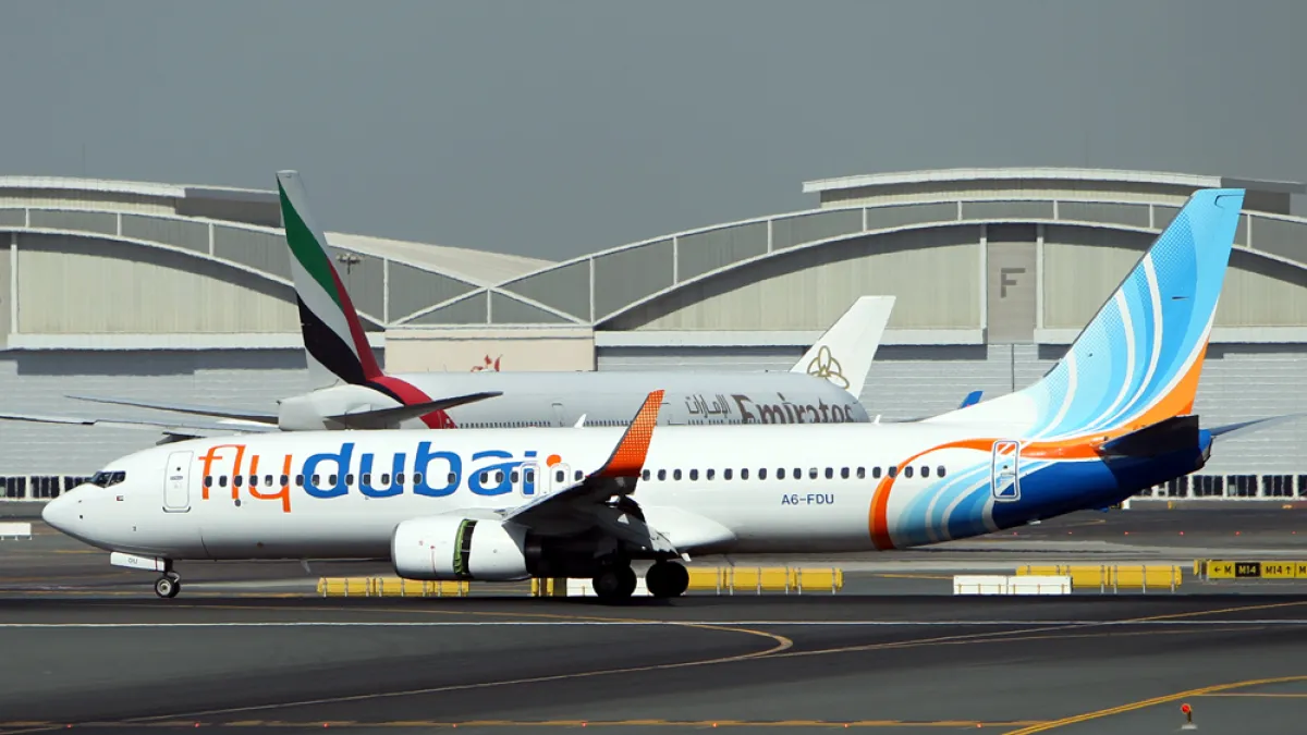A Flydubai passenger jet taxis past airport terminals, with another airliner visible behind it.