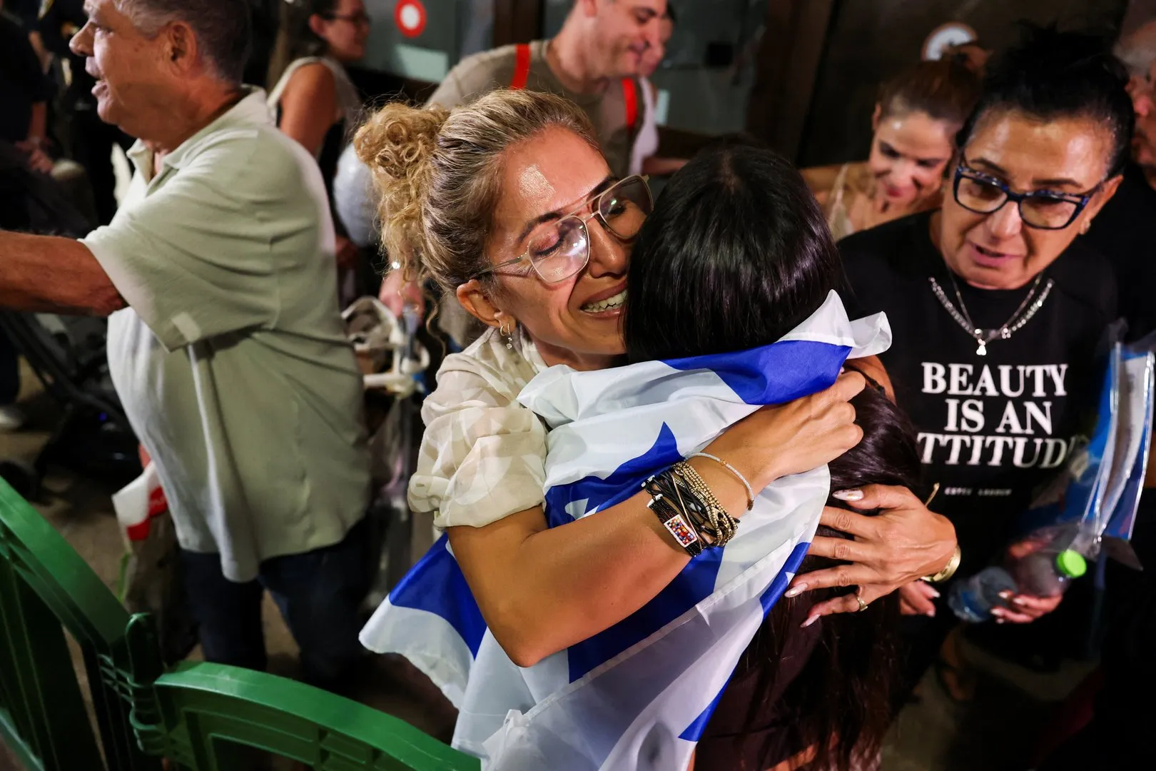 Two women embrace, one wrapped in an Israeli flag, amid a crowded indoor gathering.