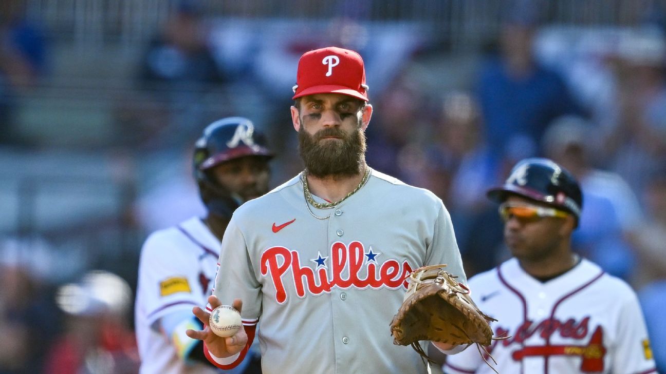 Bearded Phillies player in a gray uniform holds a baseball and glove between two Braves players.