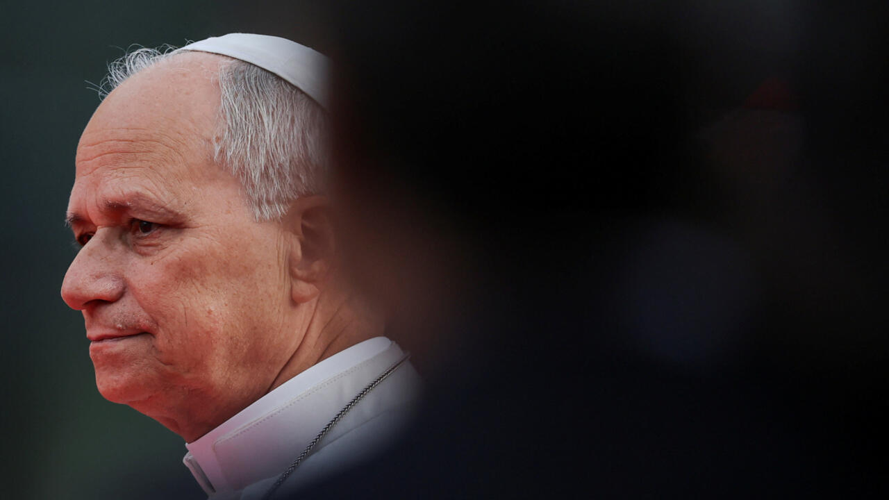 A white-haired man in white clerical clothing looks left, partially obscured by a dark foreground blur.
