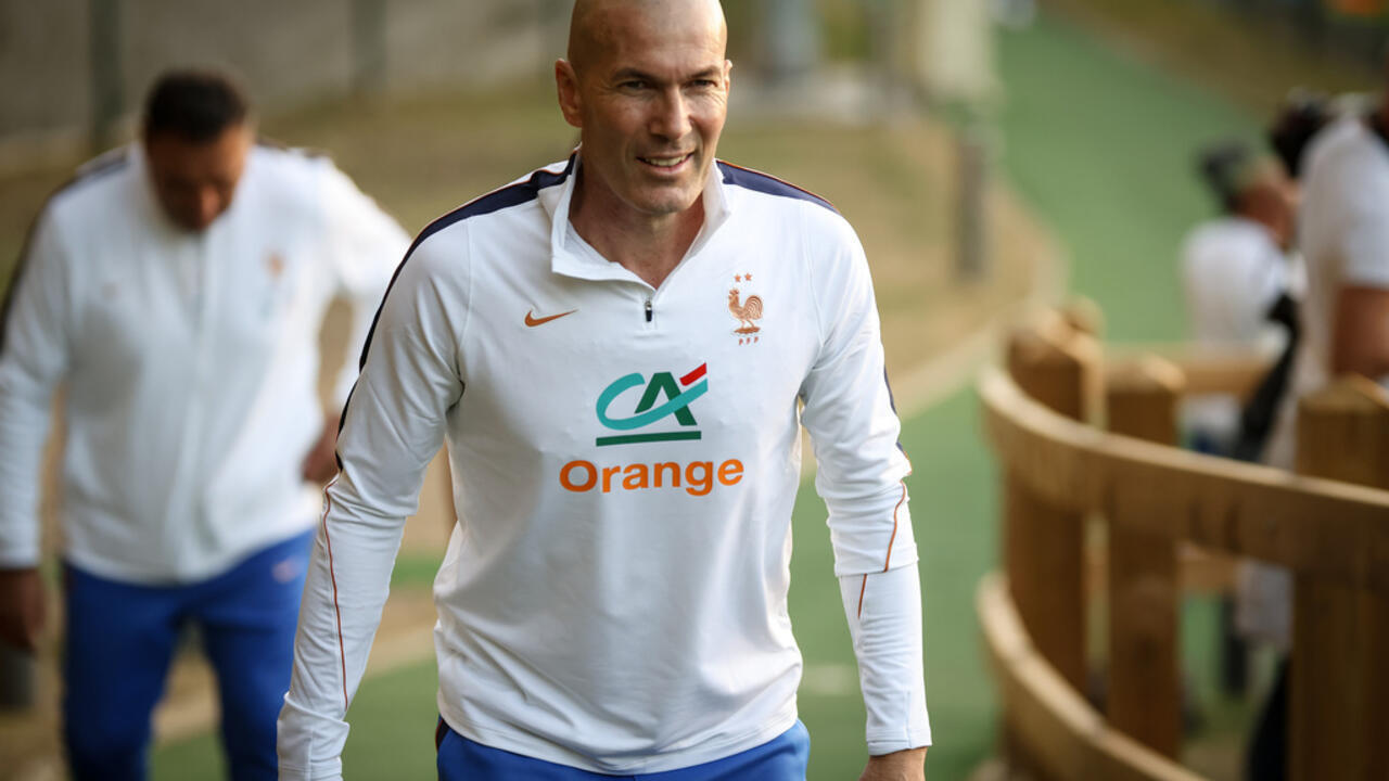 Bald football coach in white France training top walking toward camera on a practice field