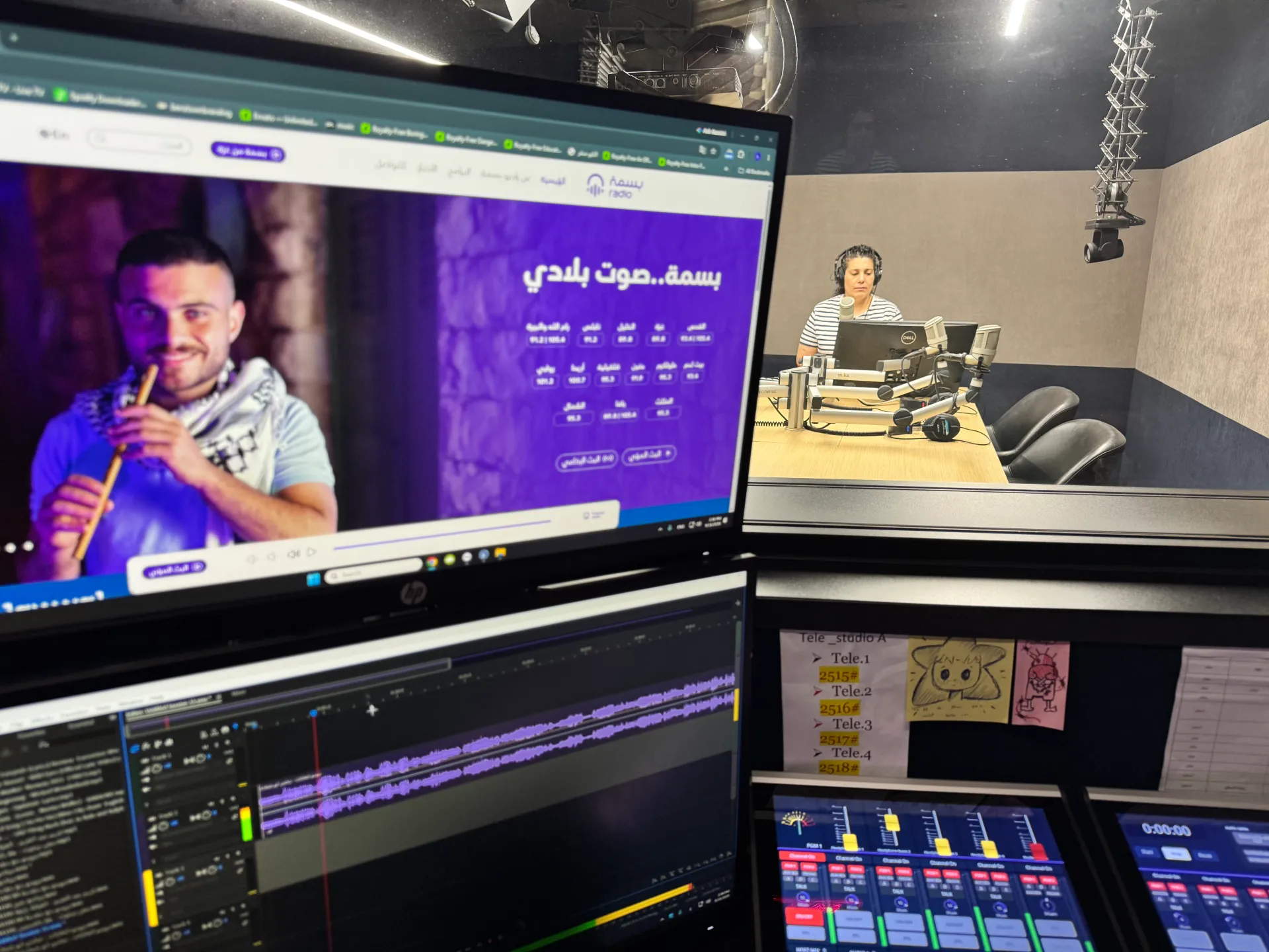 A radio studio with a woman at a console, microphones, monitors, and audio-editing software.