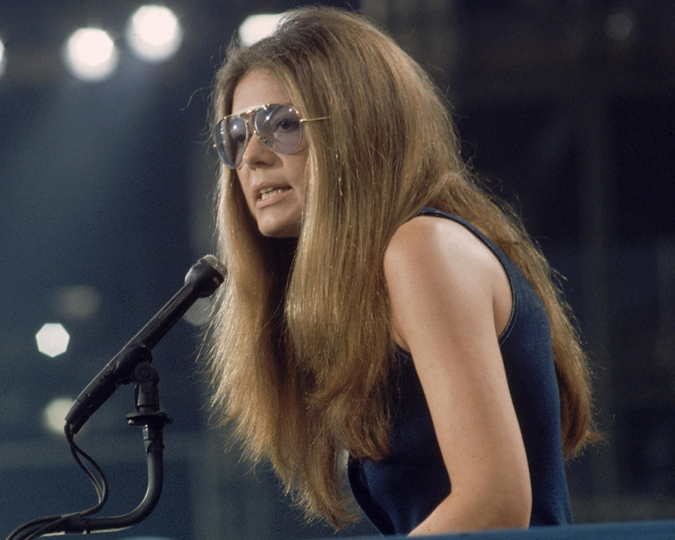Woman wearing tinted glasses speaks into a microphone during a stage performance.