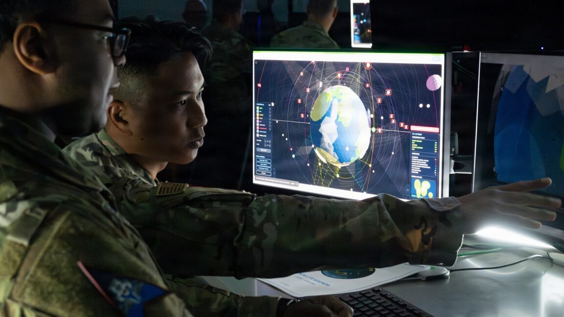 Two uniformed personnel examine an orbital tracking display showing Earth and surrounding satellites.
