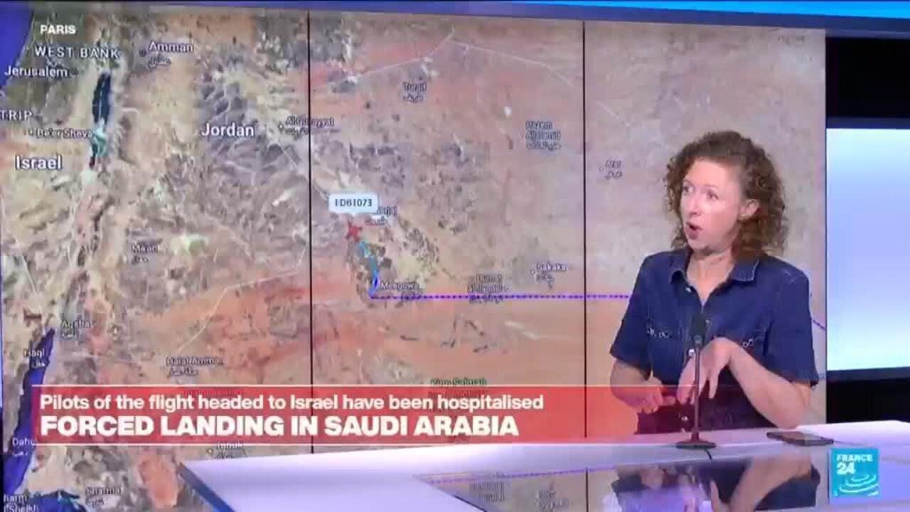 A news presenter stands beside a map as a banner reports a forced landing in Saudi Arabia.