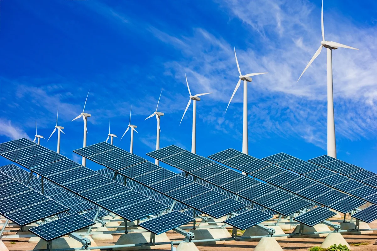 Solar panels fill foreground beneath multiple white wind turbines against a bright blue, cloud-streaked sky.