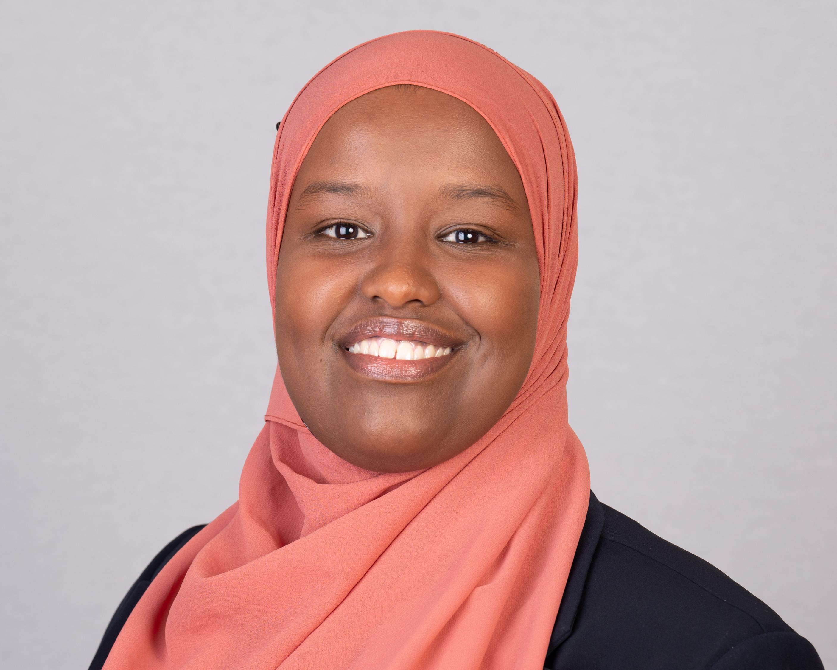 Smiling woman in a coral hijab posing against a plain grey background