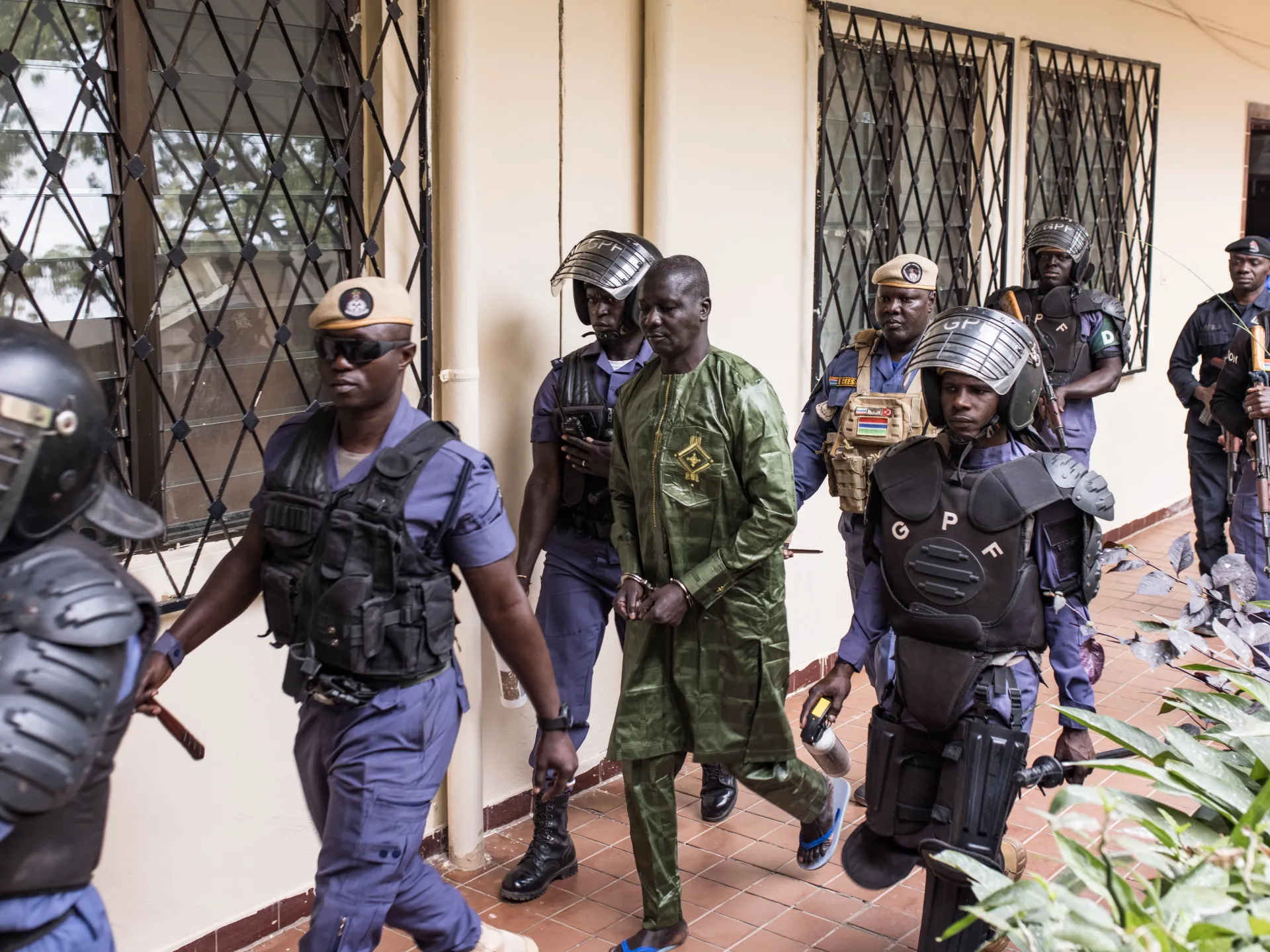 Handcuffed man in green clothing escorted by heavily armed security officers through a building