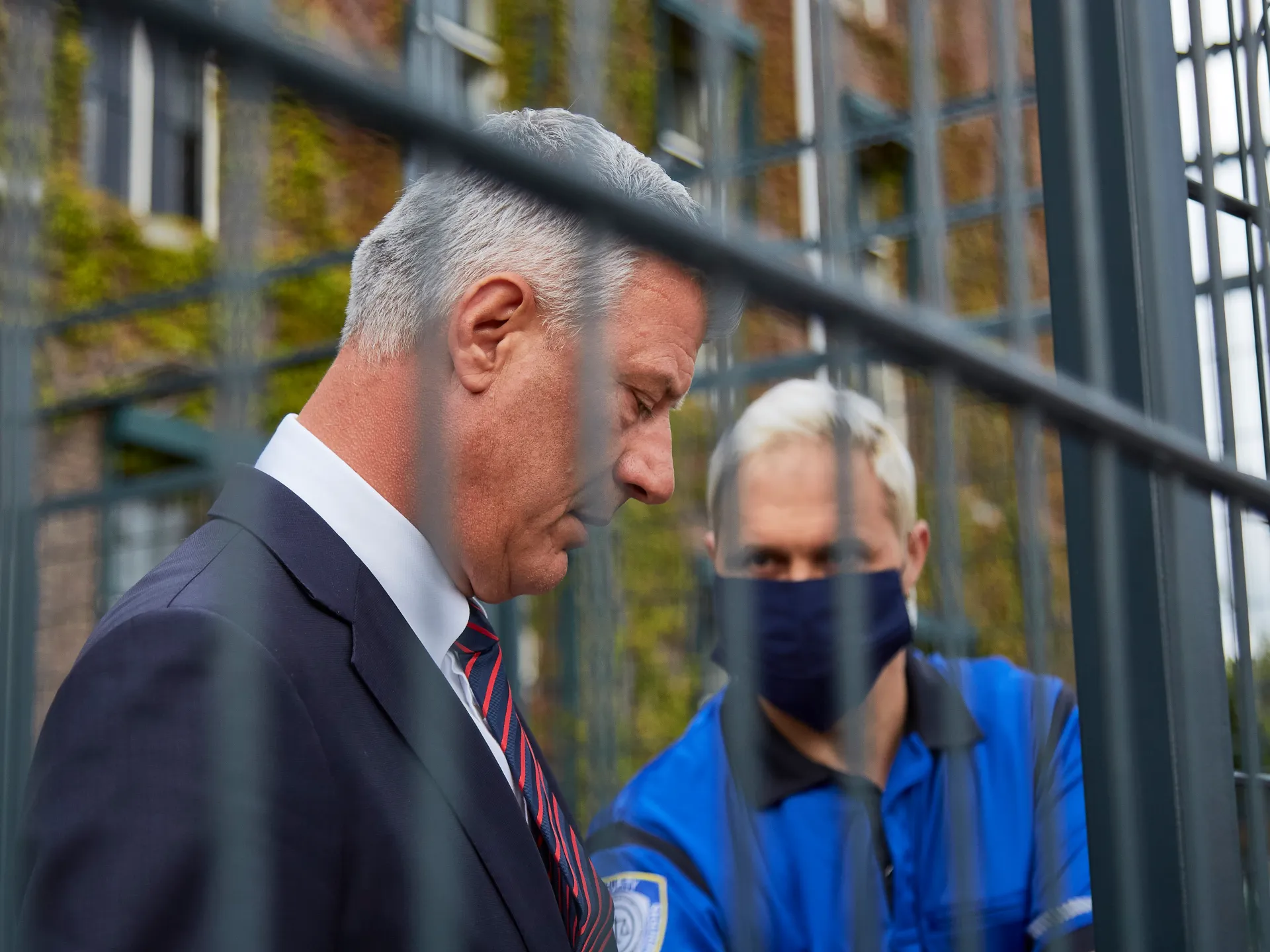 Gray-haired man in a suit stands behind metal bars beside a masked security officer.