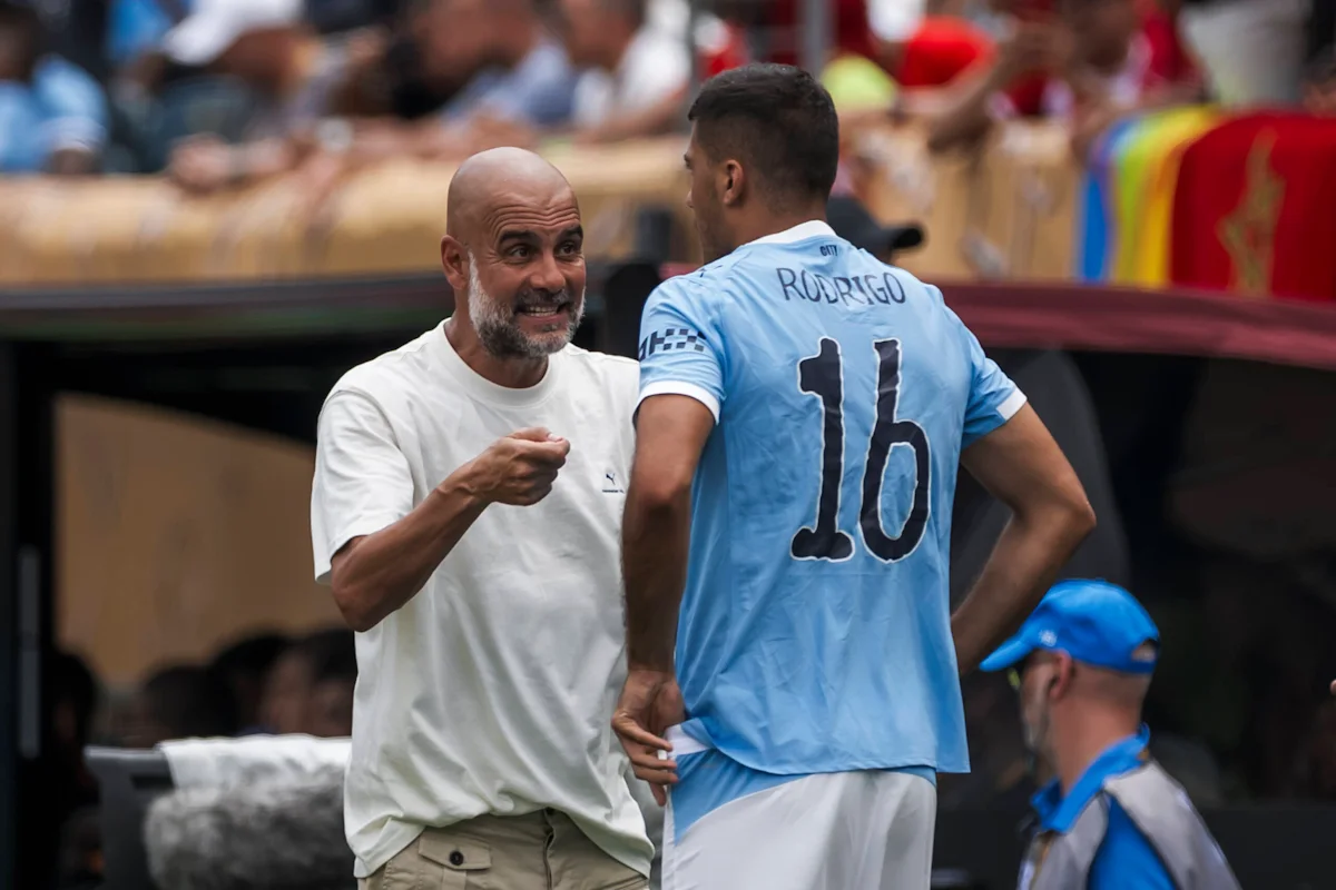 Bald coach gestures while speaking with Rodrigo, who wears a light-blue number 16 football shirt.