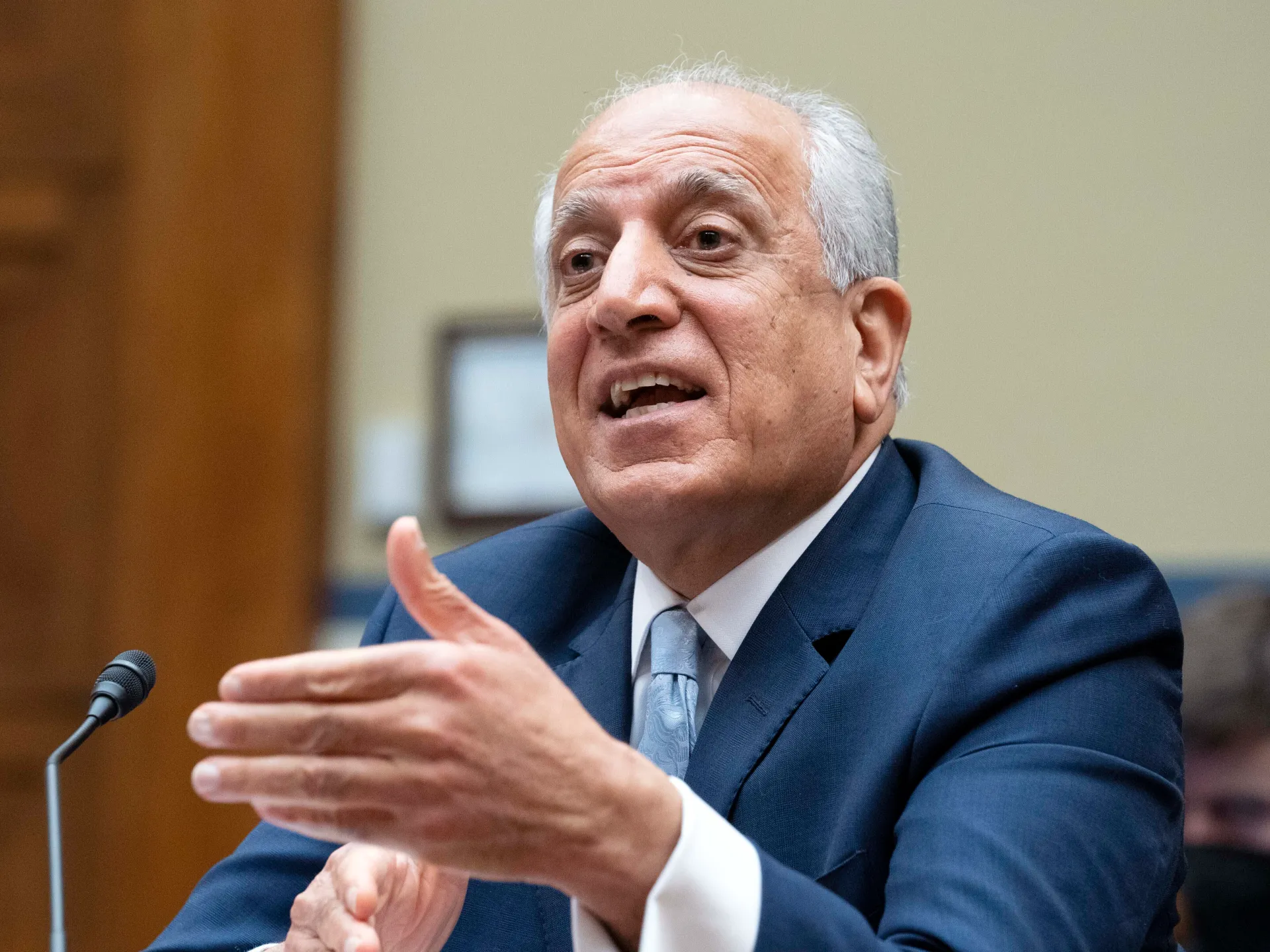 Older man in a suit gestures while speaking into a microphone at a formal hearing