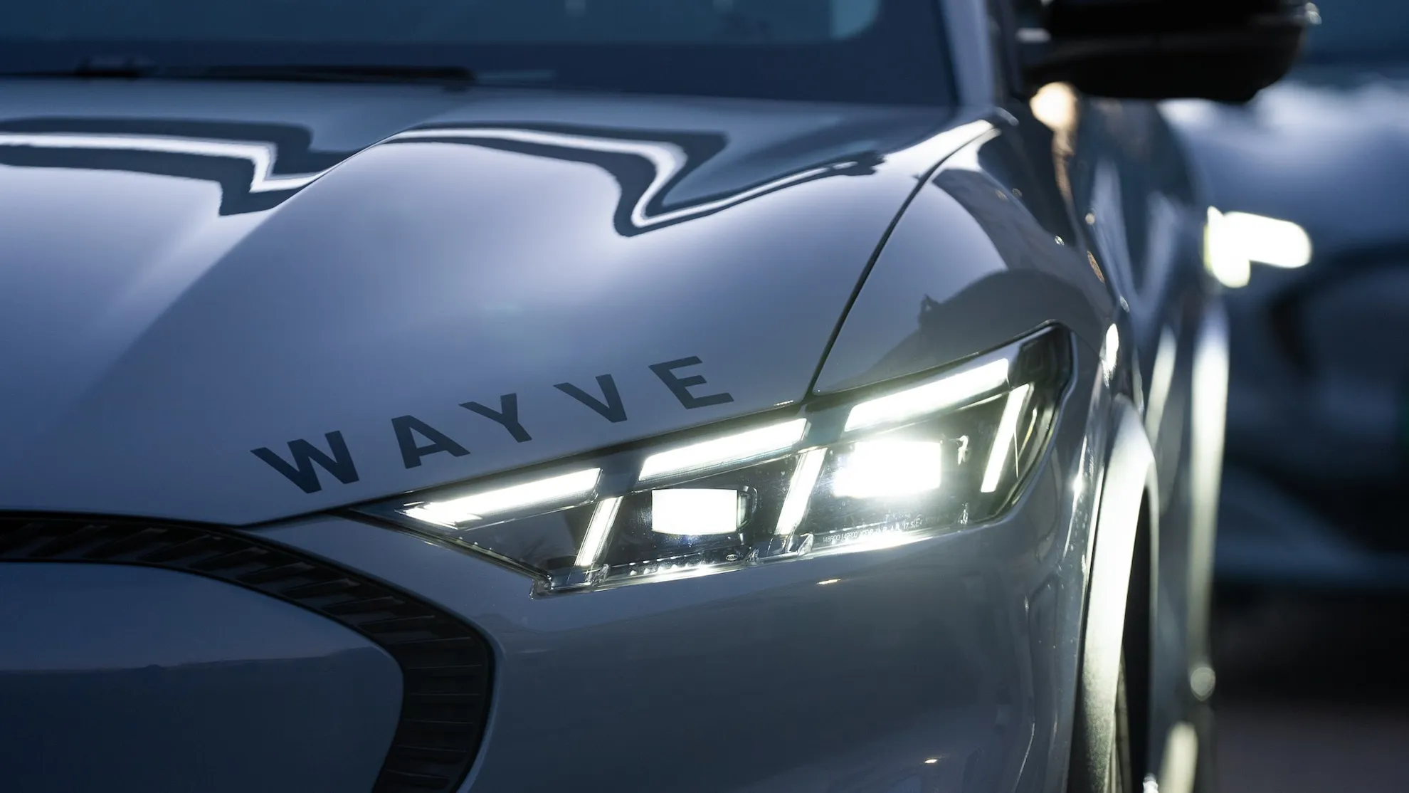 Close-up of a Wayve-branded autonomous vehicle’s front end, headlight, and hood.