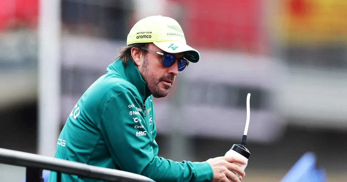 A man in a teal racing team jacket, pale cap and sunglasses holds a drink behind a trackside barrier.