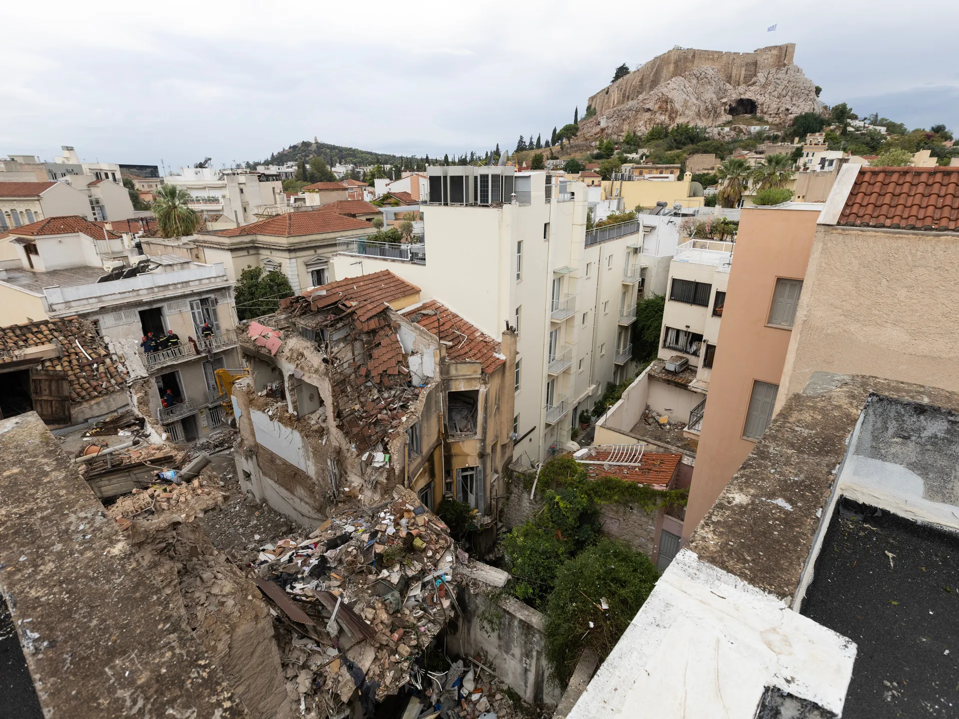 A collapsed building and rubble among Athens rooftops, with the Acropolis visible in the distance.
