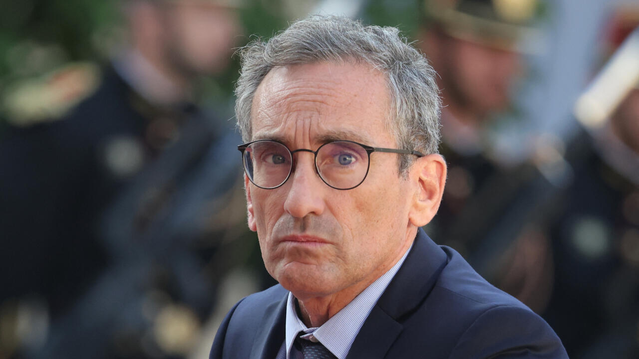 Bespectacled gray-haired man in a dark suit looking toward the camera amid a blurred crowd