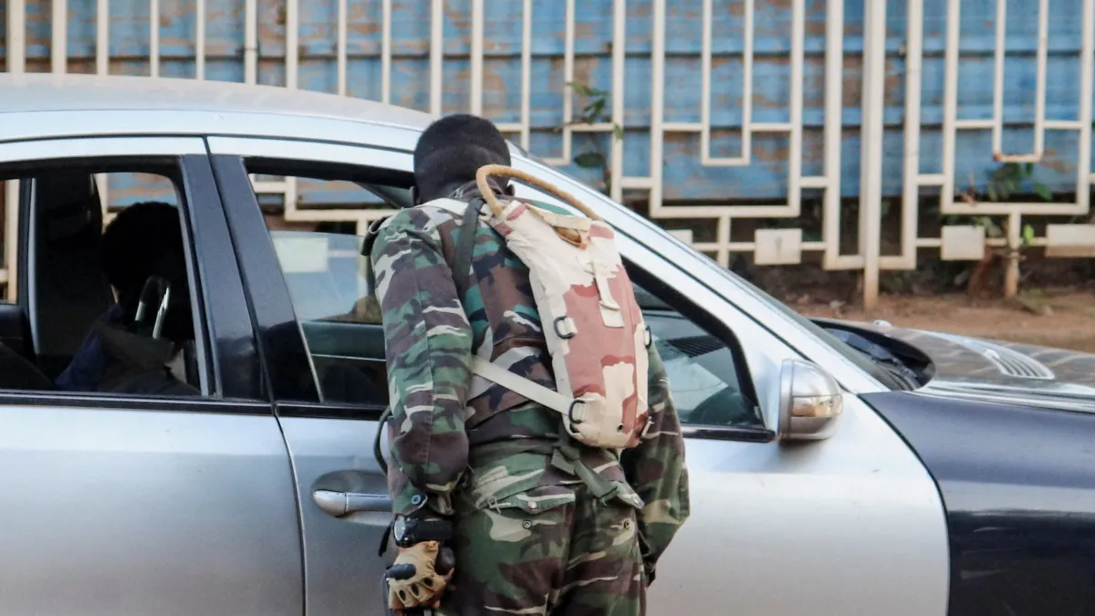 Camouflaged soldier with backpack stands beside an open white SUV outside a building.