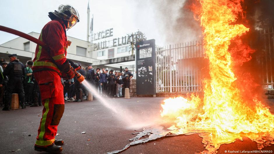 A firefighter in red protective gear sprays water toward a large street fire as a crowd watches nearby.