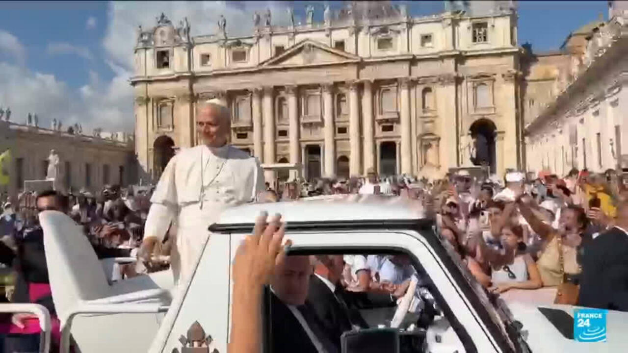 Pope-like figure rides in an open vehicle past crowds outside an ornate church