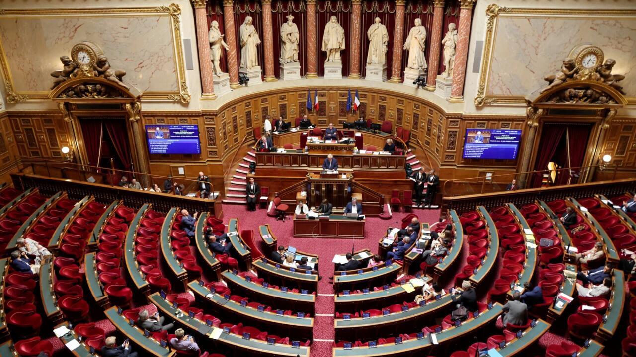 A wide view of a parliamentary chamber with curved rows of red seats and lawmakers gathered near the dais.