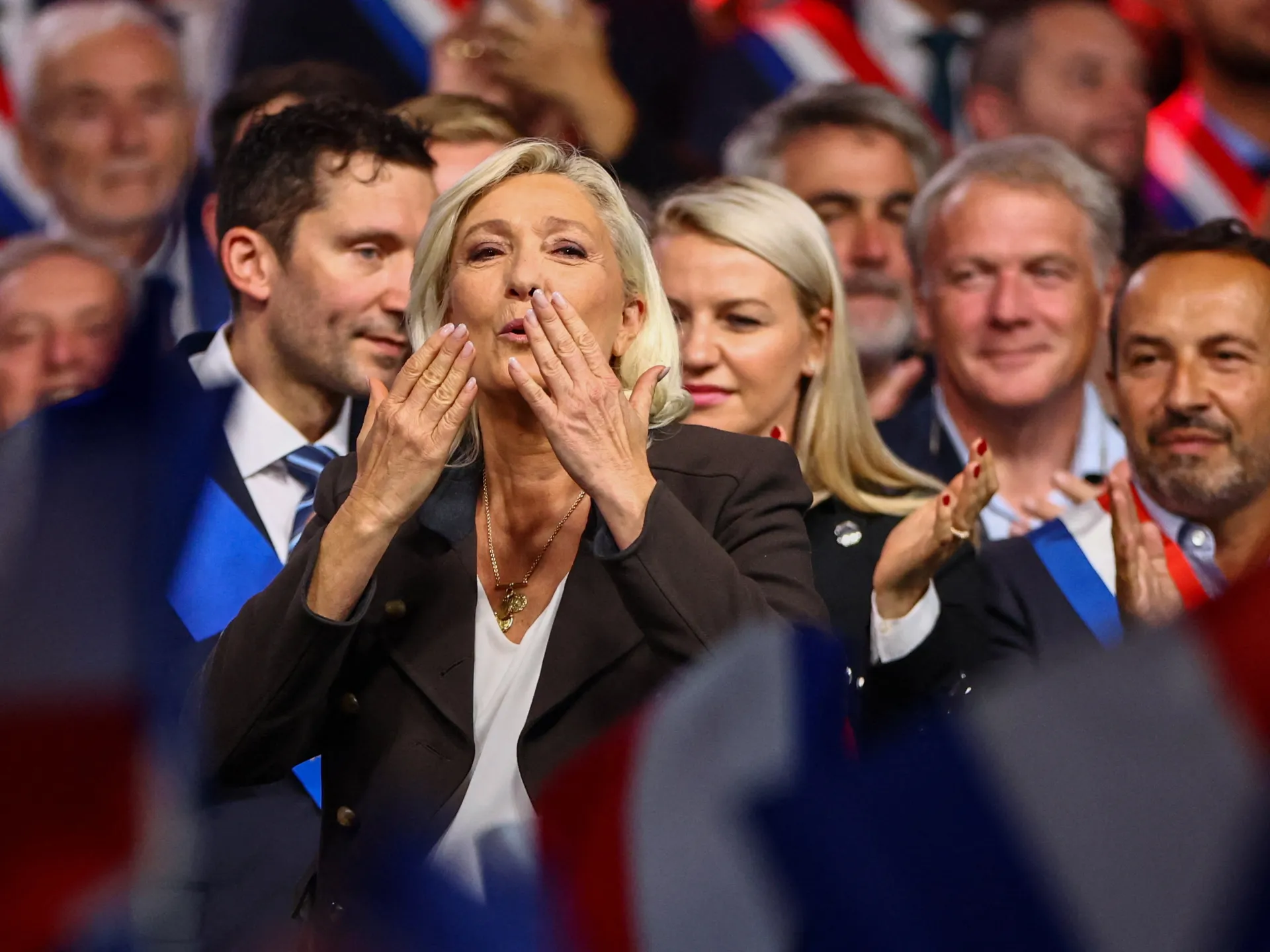 Blonde woman blows a kiss to a crowd as supporters applaud and wave French flags.