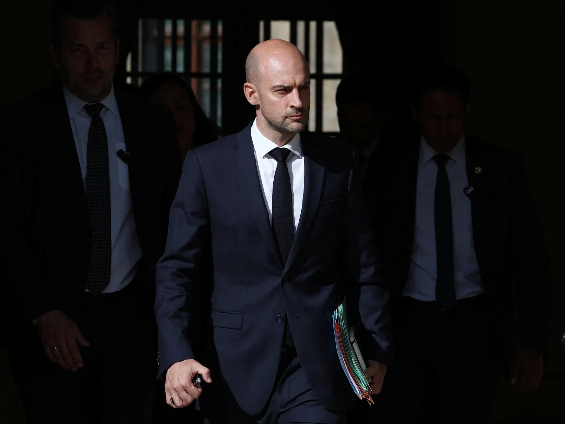 Bald man in a dark suit walks outdoors carrying colorful documents, flanked by several people.