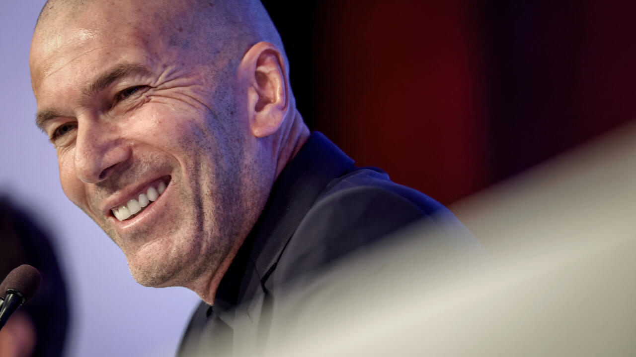 Smiling bald man in a dark jacket leaning toward a microphone during a press event