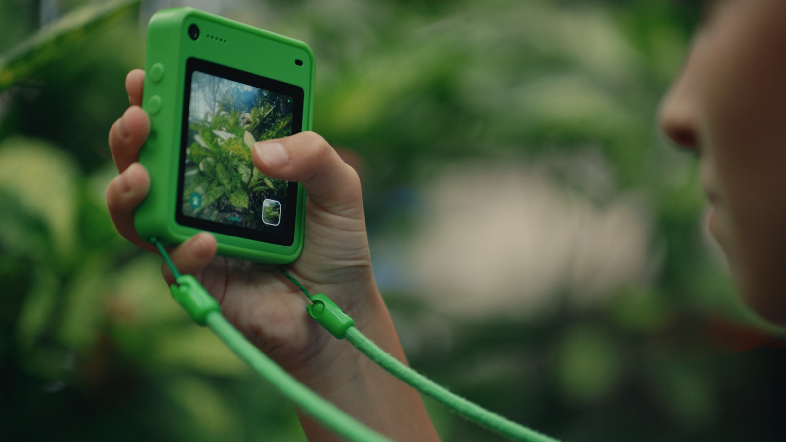 A child holds a bright green camera phone and photographs leafy plants outdoors.