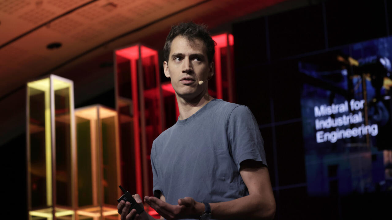 Man speaking onstage with headset microphone, holding a presentation remote in a dimly lit venue.