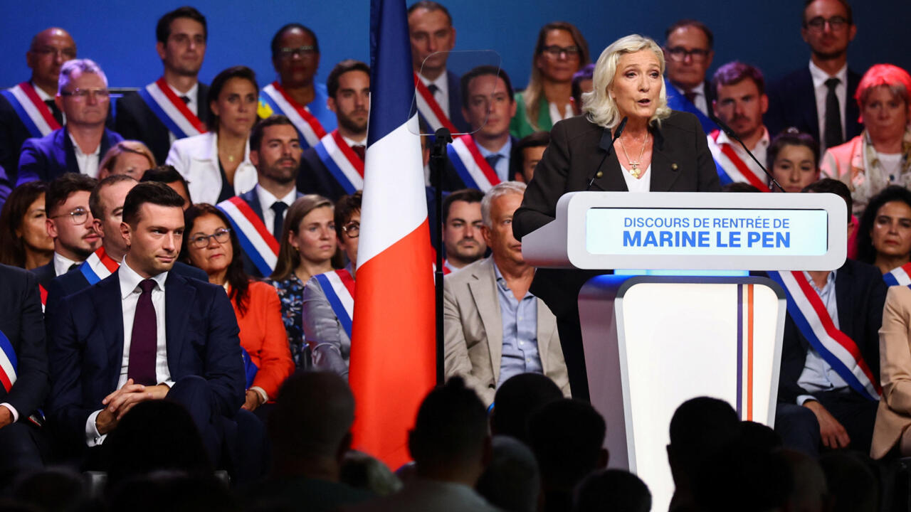 Marine Le Pen speaks at a podium beside a French flag before an audience and officials.