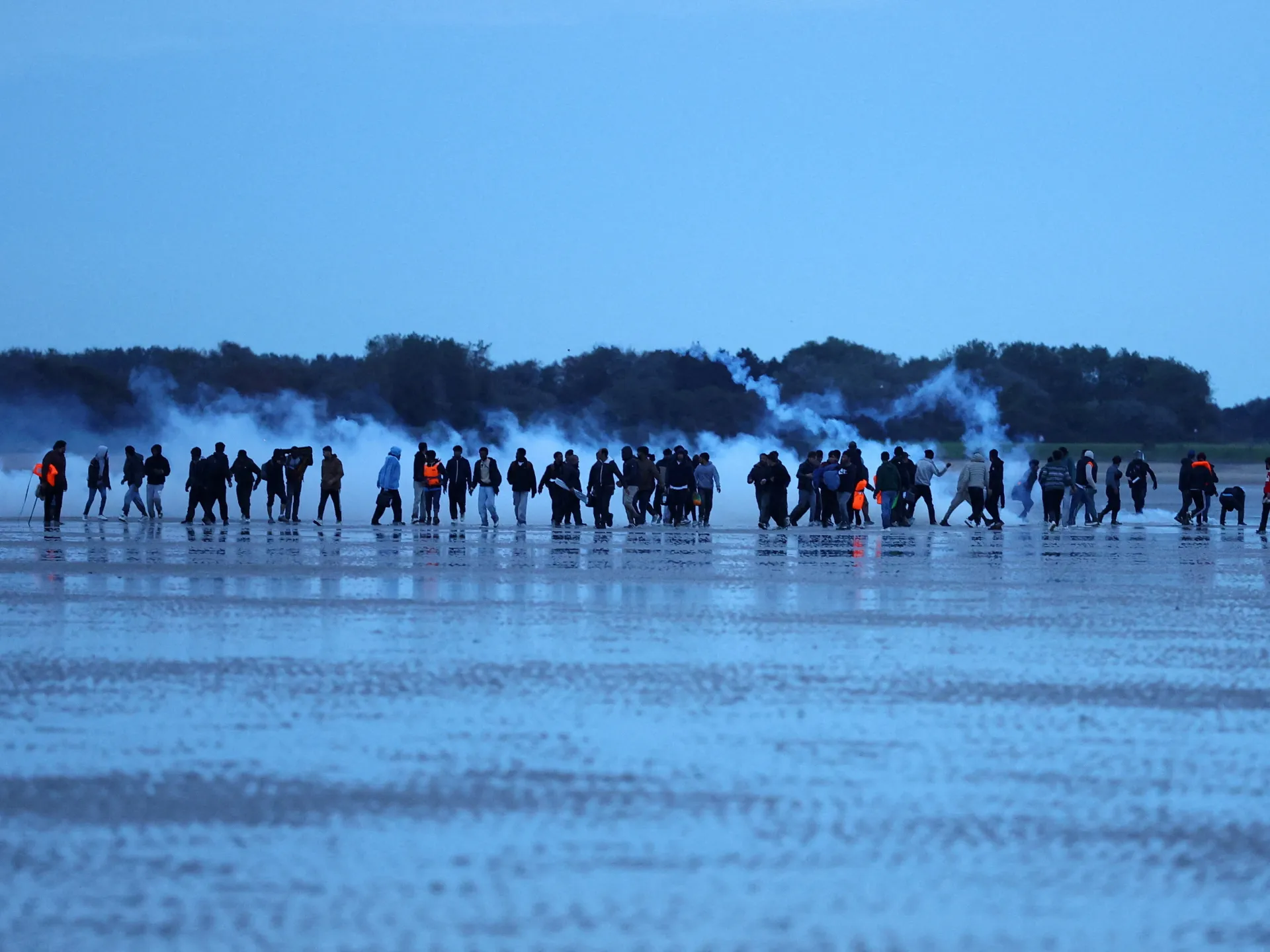 People walk across a wet shoreline as smoke rises behind them beneath a blue evening sky.