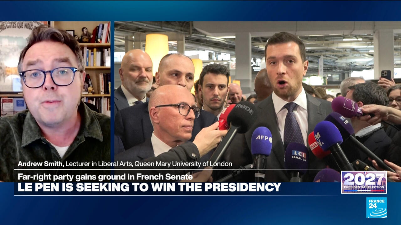 A suited man speaks to reporters amid a crowd of microphones, beside a video interview with a bespectacled commentator.