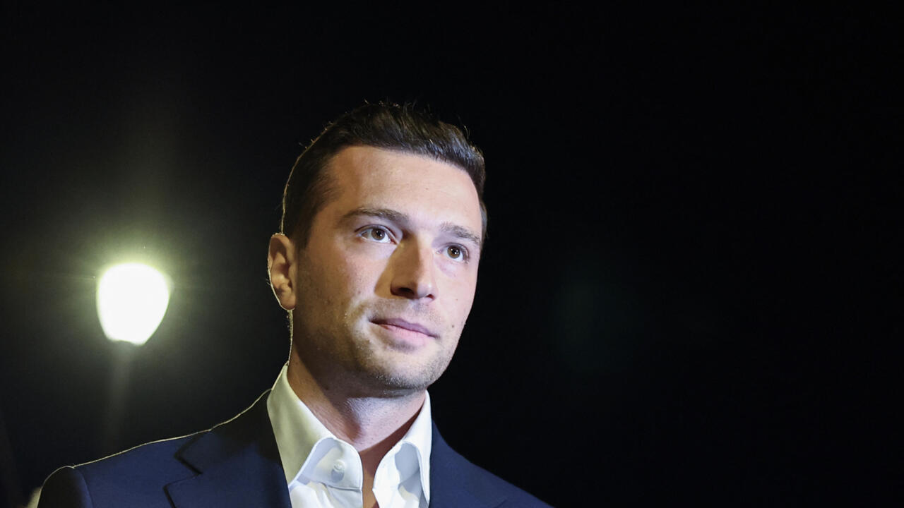A man in a dark suit and white shirt stands beneath a bright streetlight at night.