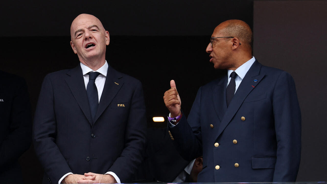 Two suited football officials stand together, one gesturing with a raised thumb during a public appearance.