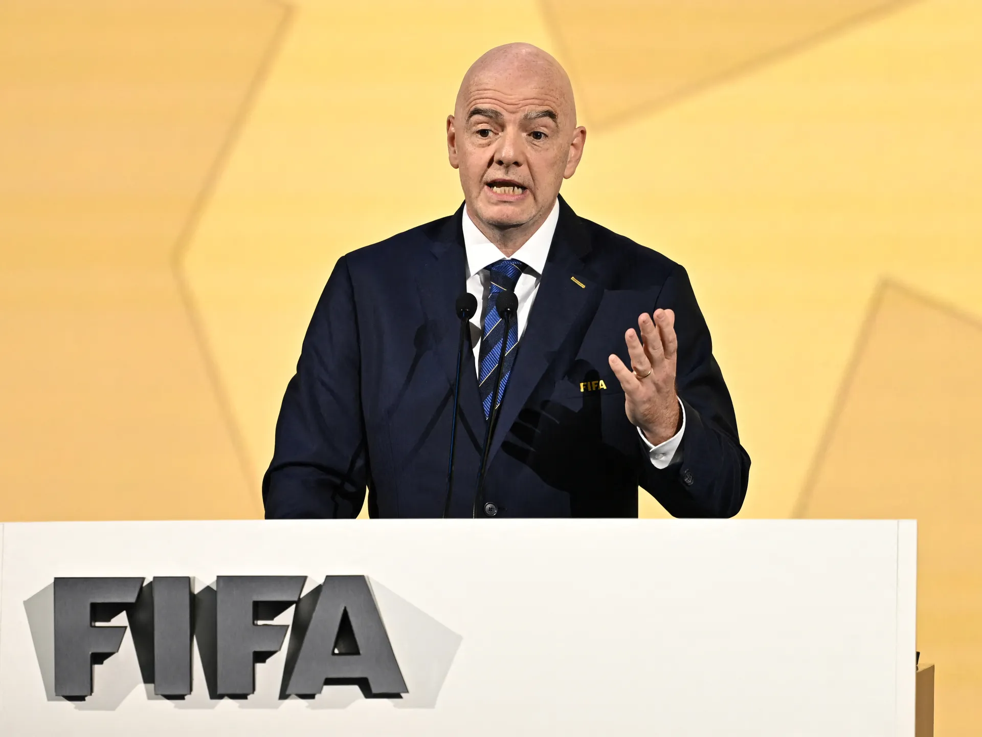 Bald man in dark suit speaking at a podium marked FIFA, gesturing with one hand.